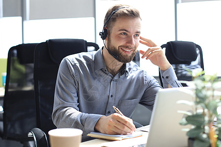 微笑的男性商业顾问戴着耳机坐在现代办公室 看着笔记本电脑屏幕进行视频通话 男士客户服务支持代理求助热线在线聊天软件服务台工作者中图片