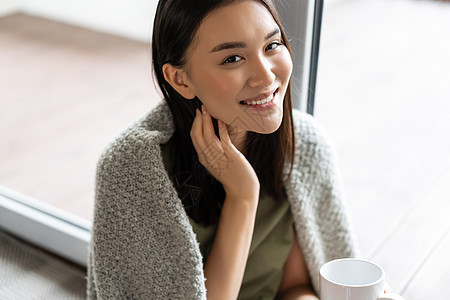 美丽的亚洲女人的画像 裹着毯子 喝着咖啡 微笑着 周末在家放松 坐在窗边欣赏美景杯子心碎疾病窗户流感疼痛鼻子保健肺炎房子图片