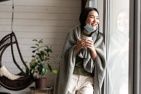 戴着医用面罩微笑的亚洲女性 在 covid 19 期间呆在家里自我隔离 生病 喝热茶 笑着看着窗外疼痛窗户感染女孩发烧肺炎疾病毯图片