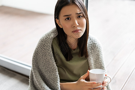 坐在窗户旁边的家中 举着咖啡杯和满脸失望 不快乐或悲哀的眼中悲伤和忧郁的女士毯子疾病肺炎女孩心碎卫生互联网杯子房子温度图片