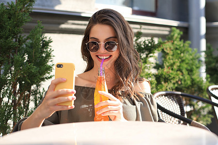 坐在街上夏日咖啡馆的美丽的黑发女人笑着 用手机拍自拍 喝果汁女士媒体休闲装微笑快乐社交连衣裙绿色城市咖啡图片