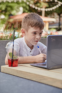 小男孩在户外用笔记本电脑做家庭作业 在复印本上写笔记童年网课咖啡店互联网会议视频学生小伙子电话小学生背景图片