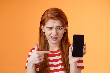 困惑失望的红发女孩展示智能手机屏幕应用程序 不确定地指着移动显示屏 完全不相信 鬼脸怀疑 犹豫 不赞成糟糕的在线选择 橙色背景女图片