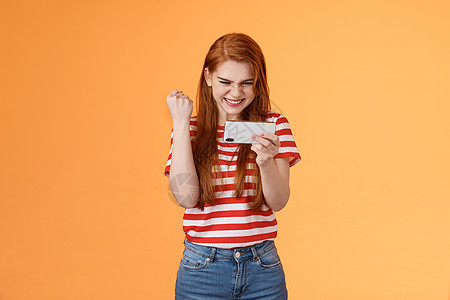 高兴的幸运红发女玩家红发 拳头满意 说是兴奋的凝视显示 水平握住智能手机 通过硬关卡玩很棒的游戏使用应用程序 取得胜利女士技术学图片