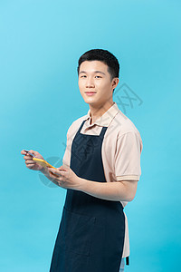 身着围裙的快乐年轻亚洲男子 在蓝背景上与世隔绝 展示了手机和信用卡职业卡片微笑食物成人人心男性店铺电话支付图片