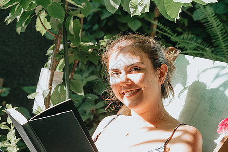 女人拿着一本书微笑着看镜头 在阳光明媚的一天倒在椅子上 复制空间 放松和爱好概念图片