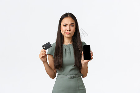 小企业主 女企业家的概念 持有信用卡的持怀疑态度和多疑的亚洲女性 在手机屏幕上显示出忧虑的情绪 白色背景售货员技术雇主促销购物金图片