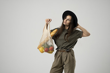 一个身穿米色 T 恤和黑色帽子的年轻时尚黑发女孩拿着一个网眼可重复使用的生态袋 里面装着食物 蔬菜和水果 零浪费 无塑料 生态友图片