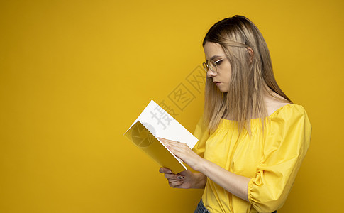 美丽聪明的年轻女孩拿着和阅读孤立在黄色背景上的书 穿着黄色衬衫 戴着眼镜看书的漂亮女人的画像 教育 学习 知识学生文学闲暇女性情图片