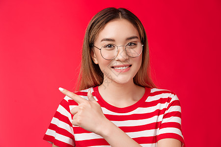 角落后面的好机会 可爱的亚洲女孩戴着眼镜条纹夏季 T 恤指向左边 微笑着露齿友好的表情 女人介绍促销表明不错的选择 红色背景快乐图片