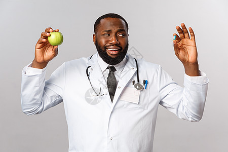 医疗保健 医学和健康的生活方式概念 持怀疑态度的英俊的非洲裔美国医生看起来不确定和犹豫 药丸显示药物和苹果 健康饮食或药物疫苗隔图片