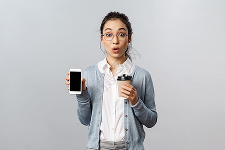 办公室生活方式 商业和人的概念 戴眼镜的亚洲女性 双唇兴奋 凝视相机 在朋友时间线上展示很棒的新应用或照片 拿着智能手机和咖啡大图片