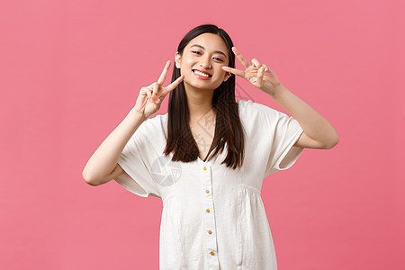 美丽 人们的情感和夏季休闲概念 傻傻而温柔的笑着 快乐的日本女孩穿着白裙 展示着和平的卡哇伊标志 笑着欢快 站着粉红色的背景图片