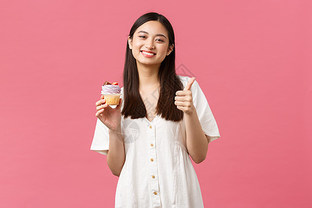 美食 咖啡和餐馆 夏季生活方式概念 快乐的亚洲女性满足于美味的甜点品味 在面包店推荐蛋糕时缩起拇指 站在粉红背景上图片