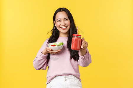 友善而愉快的笑笑的亚洲女孩伸出手来给你香水 拿着沙拉 一起吃午餐 还有黄色背景图片