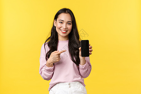 人们的情感 生活方式的休闲和美容理念 20 多岁微笑的亚洲女性 展示智能手机显示屏 推荐应用程序或手机游戏 手指指向屏幕 黄色背图片