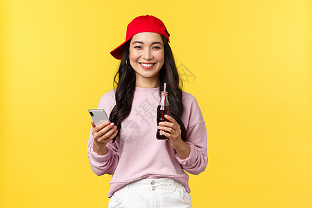 年轻的韩国女孩穿着红色帽子 发短信 使用智能手机和喝苏打饮料 站立黄色背景的喜悦 (Y L K T A )背景图片