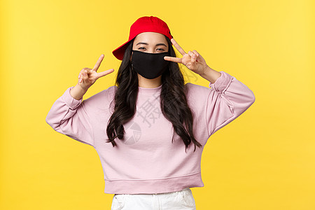 Covid-19 远离社会的生活方式 防止病毒传播的概念 戴着红帽和面罩的友善开朗的亚洲女孩 展示着和平标志 微笑着站在黄色背景图片