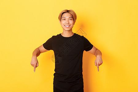 金发 穿着黑色衣服 手指向下 指着路 站在黄色背景中的微笑迷人韩国男人的肖像男朋友发型快乐职业促销教育广告工作室潮人员工图片