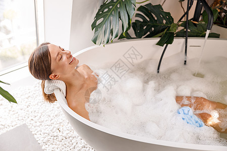 在用植物装饰的漂亮浴室里泡泡浴时 放松的女人看起来很开心图片