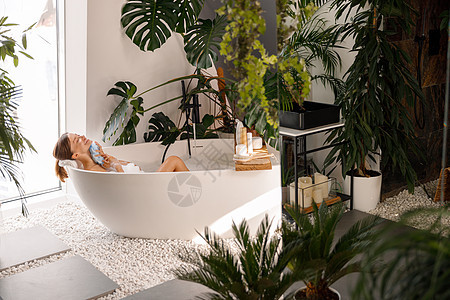 在现代浴室内与热带植物装饰的室内洗浴室里洗澡的放松的年轻女子图片
