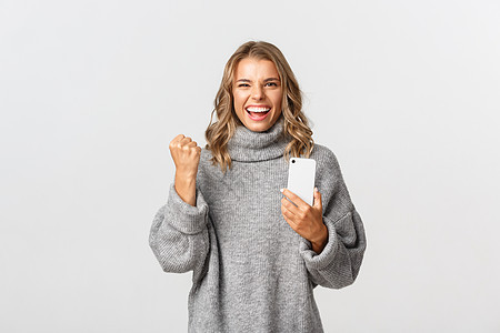 穿着灰色毛衣的快乐获胜女孩的形象 说是和胜利 拿着手机 在智能手机应用程序中实现目标 站在白色背景上互联网微笑成就情感广告购物成图片