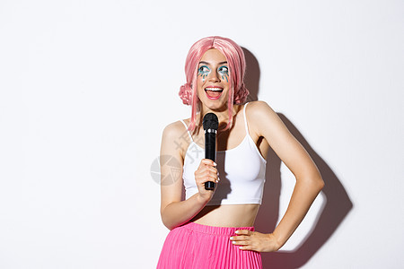 戴着粉色假发的美丽微笑女孩的形象 用麦克风唱歌 穿着万圣节服装参加派对 站在白色背景上图片