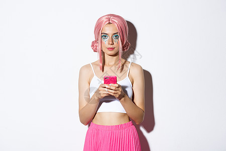穿着粉红色假发的有吸引力的派对女孩肖像 配着明亮化妆 拿着智能手机和自信地看摄影机图片