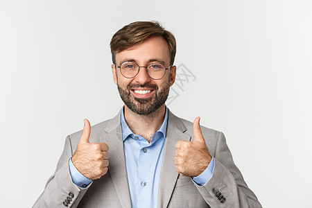 戴着眼镜和灰色西装 留着大胡子的帅哥的特写镜头 竖起大拇指表示赞同 比如推荐产品或品牌 站在白色背景上喜悦情感广告微笑快乐员工金图片