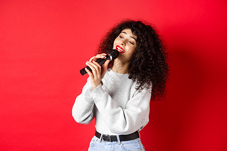娱乐和爱好概念 时尚的年轻女子唱卡拉OK 仰视并拿着麦克风 表演歌曲 站在红色背景上 笑声情感女孩歌手唱歌购物工作室魅力促销化妆图片