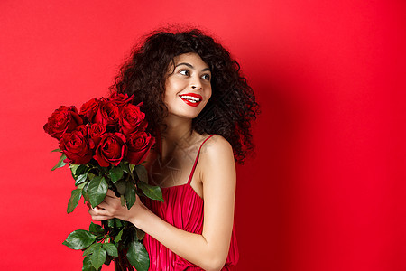 身着优雅红裙的温柔年轻女性 手捧浪漫的红玫瑰花束 看起来很正确 微笑着梦幻 在情人节想着情人 工作室背景卷曲男朋友情感盒子浪漫周图片