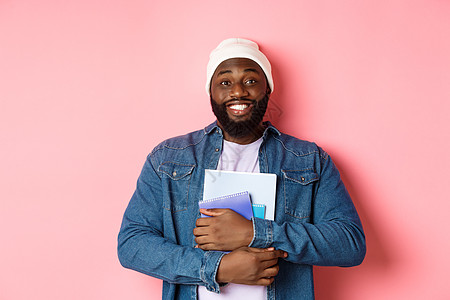 成年黑人男子持有笔记本和微笑 在课程中学习 站在粉红背景之上的图像衬衫快乐教育情感学生培训班横幅促销工作室购物图片