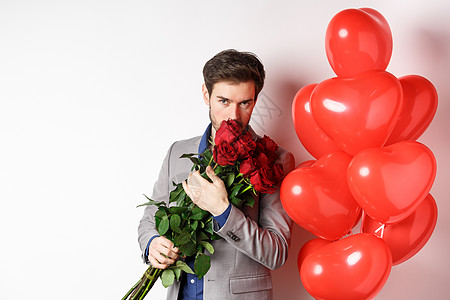 浪漫的男人闻着红玫瑰的花束 看着热辣的相机 在情人节约会上穿西装的男友与礼物和心气球 白色背景感情惊喜快乐男朋友男性展示工作室花图片