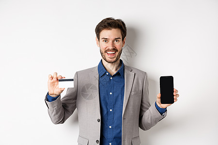 网上购物 微笑的商务人士穿着用空智能手机屏幕显示塑料信用卡 站在白色背景下站立着的牌子上企业家成人工作室情绪男性经理广告购物工人图片