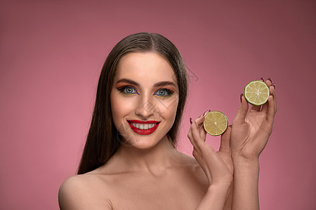 快乐的年轻女人展示了石灰对健康的好处 手里拿着两片切片 看起来很迷人 粉红色背景中长发的迷人快乐风趣女士微笑饮食眼睛食物乐趣水果图片
