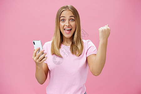女孩在智能手机游戏中打破了自己的记录 举起握紧的拳头欢呼 胜利地拿着手机 微笑着兴奋而快乐地对着镜头微笑 在粉红色背景下用快乐的图片