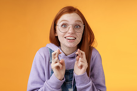 女孩兴奋地看着结果交叉手指祝你好运欢呼最喜欢的球队想赢梦想成真激动张大嘴巴凝视相机充满希望的祈求欲望实现 橙色背景女士青少年学生图片