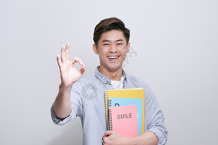 背着背包笔记本的年轻学生男子在孤立的白色背景上开心地笑着做 ok 的手势 用手指竖起大拇指 优秀的标志家伙高清图片素材