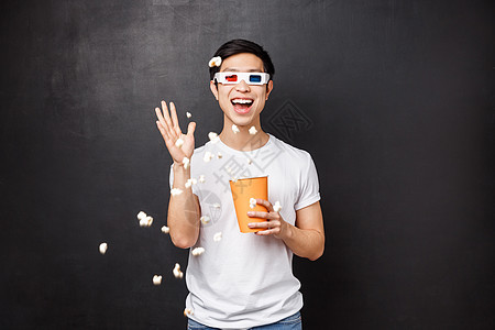 休闲 电影和生活方式的概念 穿着 T 恤的有趣而无忧无虑的风趣亚洲男人在电影院屏幕上扔爆米花 一边看电影 3D 眼镜 一边站在黑图片