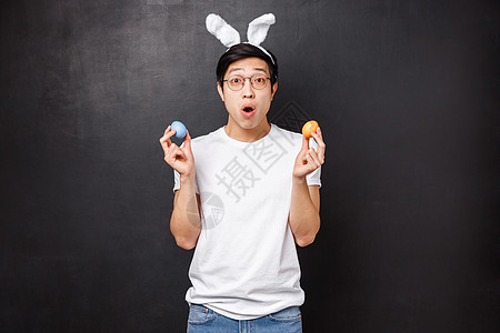 假期 聚会和复活节的概念 戴着兔子耳朵的亚洲年轻人惊讶地拿着彩绘鸡蛋作为学习女朋友的传统 张开嘴着迷于了解新文化 黑色背景图片