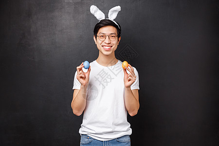 假期 聚会和复活节的概念 戴着兔子耳朵庆祝正统圣日的快乐微笑友善亚洲人的肖像 拿着彩蛋 看着相机 站在黑色背景上图片