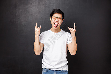 戴着眼镜和 T 恤的快乐兴奋和外向的亚洲男人 玩得开心展示舌头闭上眼睛放松 并在听很棒的歌曲 黑色背景时做出摇滚重金属手势图片