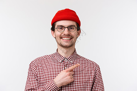 特写肖像帅气友善的嬉皮士男性 戴着红色豆帽和眼镜 微笑着满足和快乐 指着右上角 展示很酷的产品或地方闲逛图片