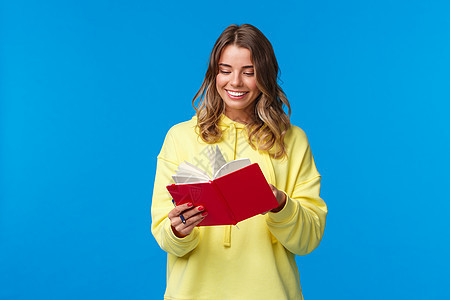 女孩买了新的计划书 写下笔记或演讲 开心地微笑 拿着红色笔记本 决定开始秘密日记 站在蓝色背景的黄色连帽衫家庭作业快乐幸福金发女背景图片