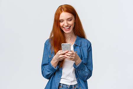 聊天 交流和移动概念 快乐的红头笑脸女孩发短信朋友 笑得像看滑稽Vlog一样 看着智能手机显示高兴 站立白背景图片