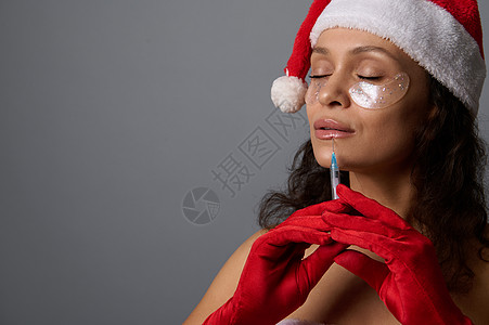 美丽的女人穿着圣诞老人的嘉年华装束 眼睛下方有斑块 嘴唇上正在接受美容注射 美容美容院圣诞广告的丰唇概念图片