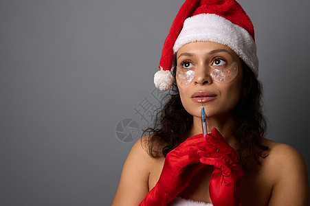 美丽的女人 有着自然的妆容和眼睛下面的补丁 戴着圣诞老人的帽子 拿着注射器 靠近她的脸 看着灰色背景的复制空间 注射美容概念图片