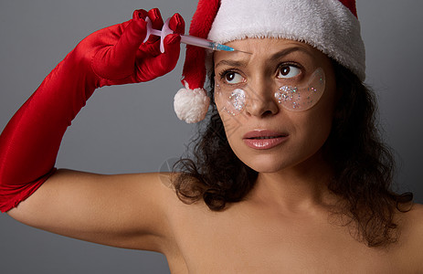 戴着圣诞帽和红色手套的漂亮女人拿着小注射器 在额头上做美容注射 抗衰老治疗 水疗沙龙圣诞广告的复兴概念图片