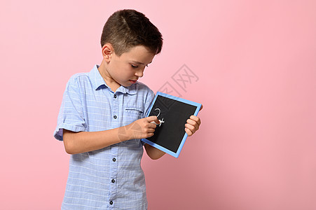 一个穿蓝衬衫的男孩在黑板上写着粉笔 与粉红背景隔绝 复制空间 回到学校 概念图片