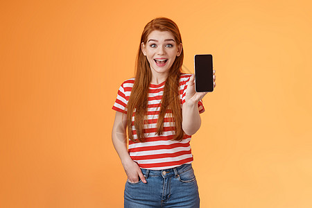 兴奋可爱外向的红发女性给展示应用留下深刻印象 拿着智能手机介绍小工具功能 微笑惊讶 看起来像应用一样热情 吹嘘游戏分数 橙色背景图片